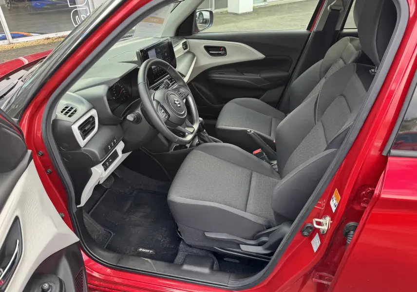 Intérieur avant gauche de la Suzuki Swift 1.2 Hybrid Privilege 2025 en rouge Burning Pearl, sièges gris et tableau de bord moderne.