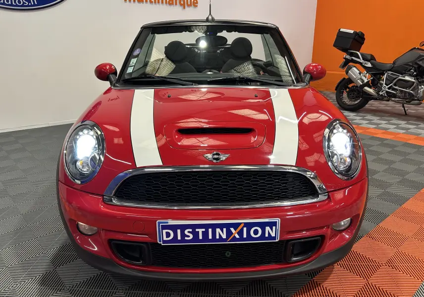 Vue frontale d'une MINI Cabriolet Cooper S rouge avec bandes blanches sur le capot et phares allumés dans un showroom.