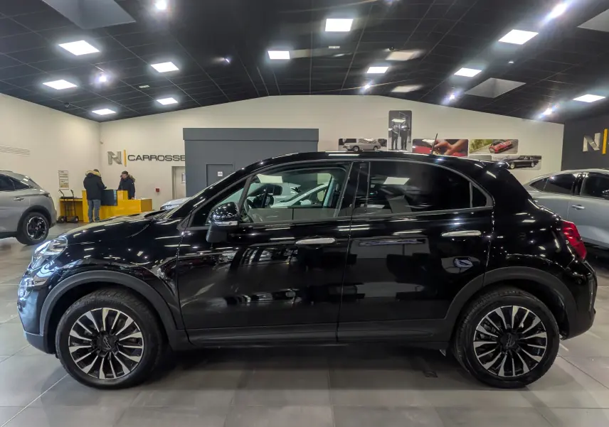 Vue de profil côté gauche d'une FIAT 500X 2024 noire Cinéma avec jantes alliage bicolores en intérieur showroom.