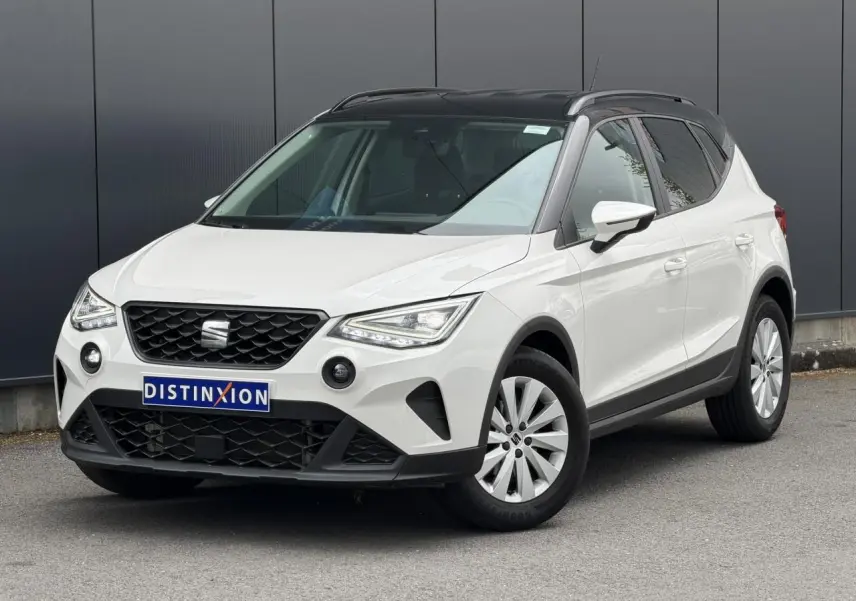 Vue 3/4 avant droite d'un SEAT Arona blanc avec toit noir et jantes alliage 16 pouces, garé devant un mur gris.