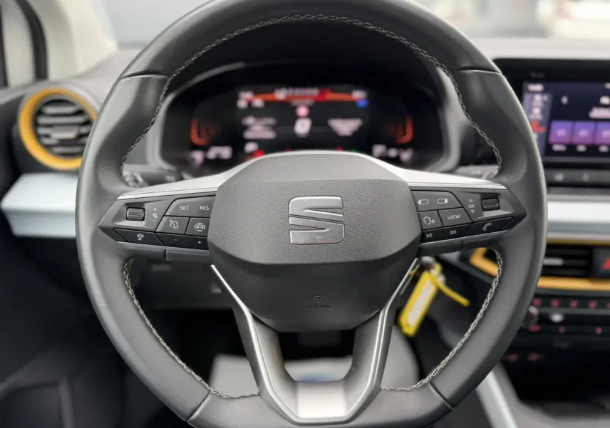 Vue rapprochée du volant noir de la SEAT Arona TSI 115 DSG Style avec commandes intégrées et tableau de bord digital flou.