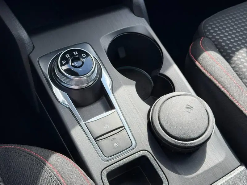 Gros plan sur la console centrale grise avec molette de boîte auto et porte-gobelets, sièges tissu noir avec surpiqûres rouges.