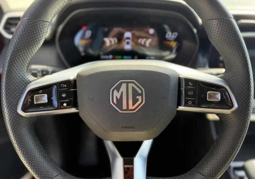 Vue rapprochée du volant en cuir noir perforé avec logo MG au centre, tableau de bord numérique en arrière-plan flou.