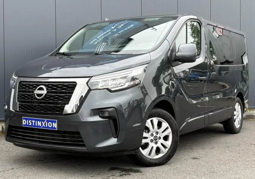 Vue 3/4 avant d’un Nissan Primastar gris comète avec hayon vitré et jantes alliage, stationné devant un mur gris.