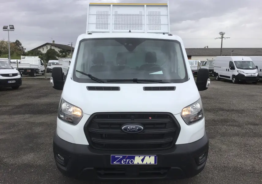 Vue frontale d'un Ford Transit Chassis Cabine blanc avec benne acier et coffre, phares allumés sur parking.