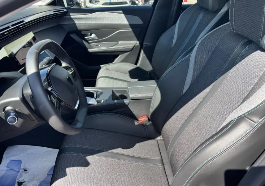 Vue intérieure côté conducteur de la Peugeot 408 Hybrid 2025 noire, avec sièges tissu et cuir, accoudoir central et volant multifonction.