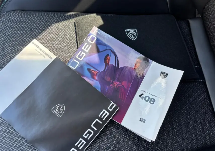 Vue rapprochée des brochures Peugeot 408 posées sur un siège tissu noir à motifs, avec logo Peugeot visible.