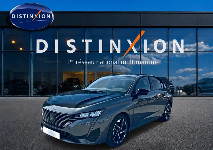 Peugeot 308 gris 3/4 avant droit, jantes alu noires, devant vitrine Distinxion avec reflet ciel bleu.