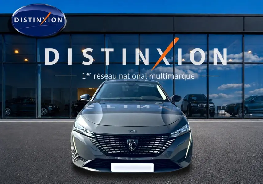 Peugeot 308 gris 3/4 avant face, avec calandre distinctive et phares LED allumés devant showroom Distinxion.