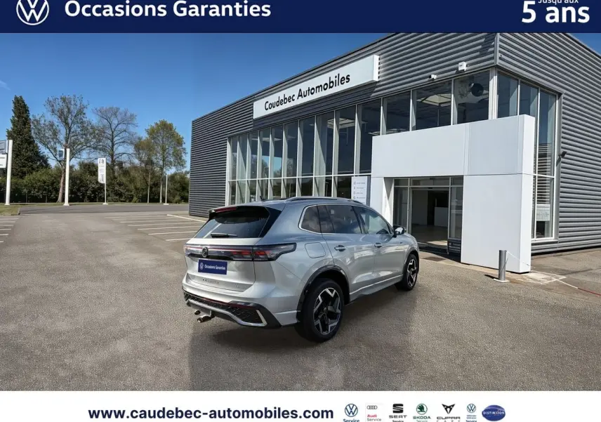Vue 3/4 arrière droite d’un Volkswagen Tiguan eHybrid argent Dolomite avec jantes 19 pouces devant un showroom Caudebec Automobiles.
