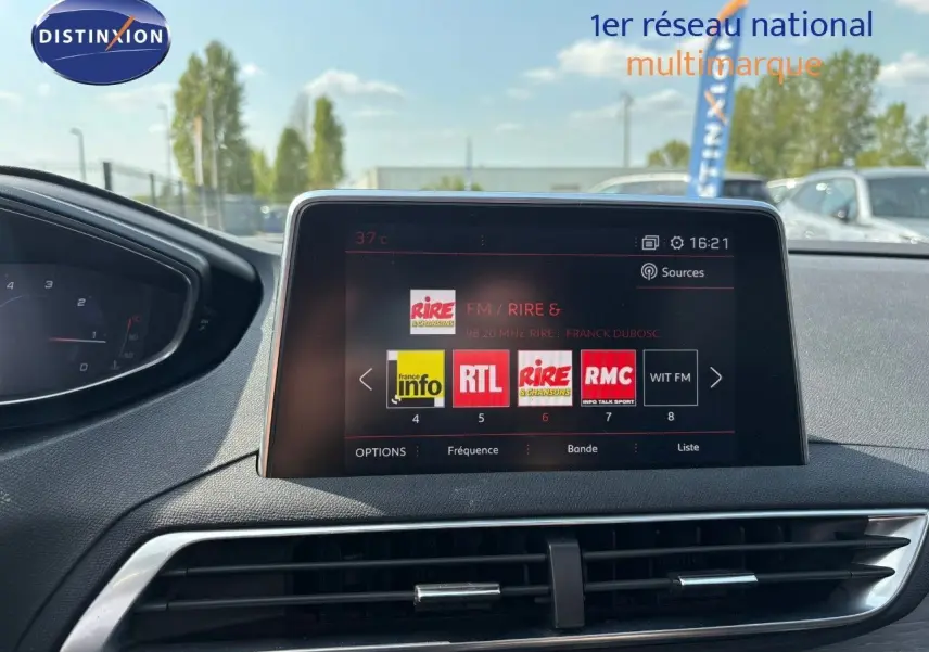 Écran multimédia central du Peugeot 3008 GT blanc nacré, avec tableau de bord partiel et vue extérieure floutée.