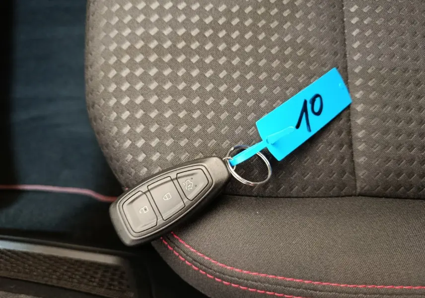 Clé de démarrage Ford Kuga 2024 posée sur siège tissu noir avec surpiqûres rouges, vue en plongée rapprochée.