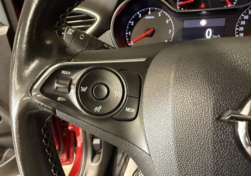 Gros plan sur le volant noir de l’Opel Crossland rouge, avec commandes de régulateur de vitesse et tableau de bord en arrière-plan.