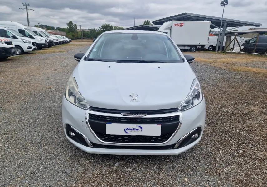 Vue avant d'une Peugeot 208 blanc nacré 2018, avec calandre chromée et phares allumés sur un sol gravillonné.