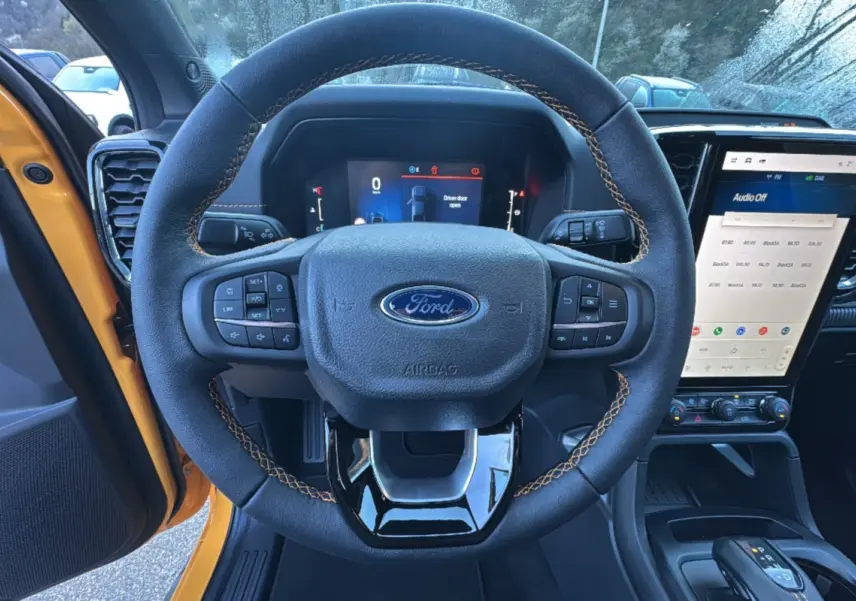 Vue intérieure centrée sur le volant cuir noir avec surpiqûres orange du Ford Ranger Cyber orange, écran tactile et tableau de bord numériques visibles.