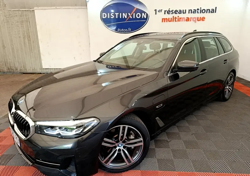 BMW Série 5 Touring 530e gris clair vue 3/4 avant droit, avec jantes alliage et phares LED allumés.