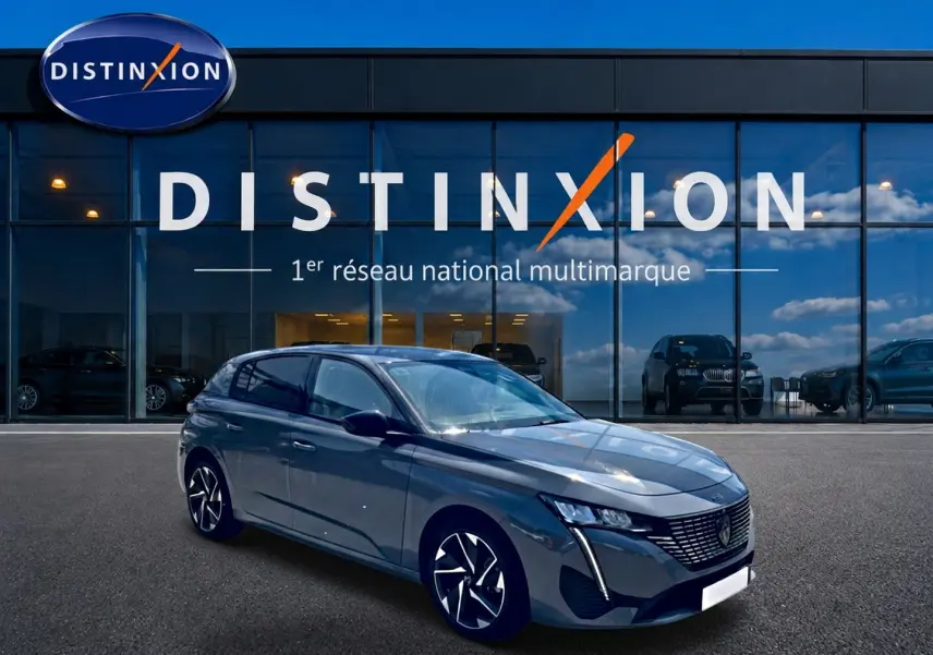 Peugeot 308 gris 3/4 avant droit, jantes alu noires et calandre distinctive, devant vitrine Distinxion.