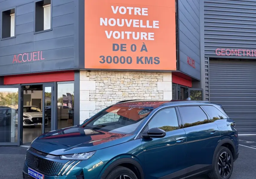 Peugeot 5008 hybride vert en 3/4 avant droit, avec jantes noires et calandre distinctive, devant un bâtiment commercial.