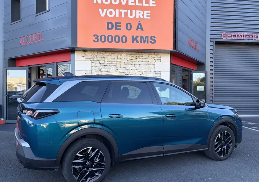 Vue côté gauche d’un Peugeot 5008 hybride vert 2025, avec jantes noires et toit noir, stationné devant un bâtiment commercial.