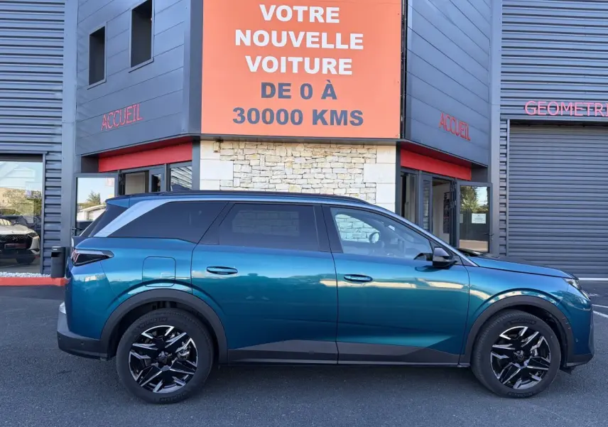 Profil côté gauche d’un Peugeot 5008 hybride vert métallisé, garé devant un bâtiment avec panneau orange.