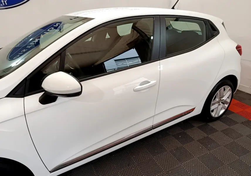 Profil côté gauche d'une Renault Clio Business TCe 100 blanche avec rétroviseur et poignées assortis, sur sol damier noir et rouge.