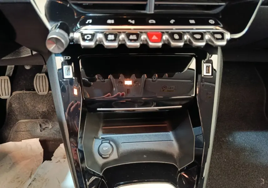 Vue rapprochée de la console centrale noire brillante de la Peugeot 208 BlueHDi 100, avec boutons et prise 12V visible.