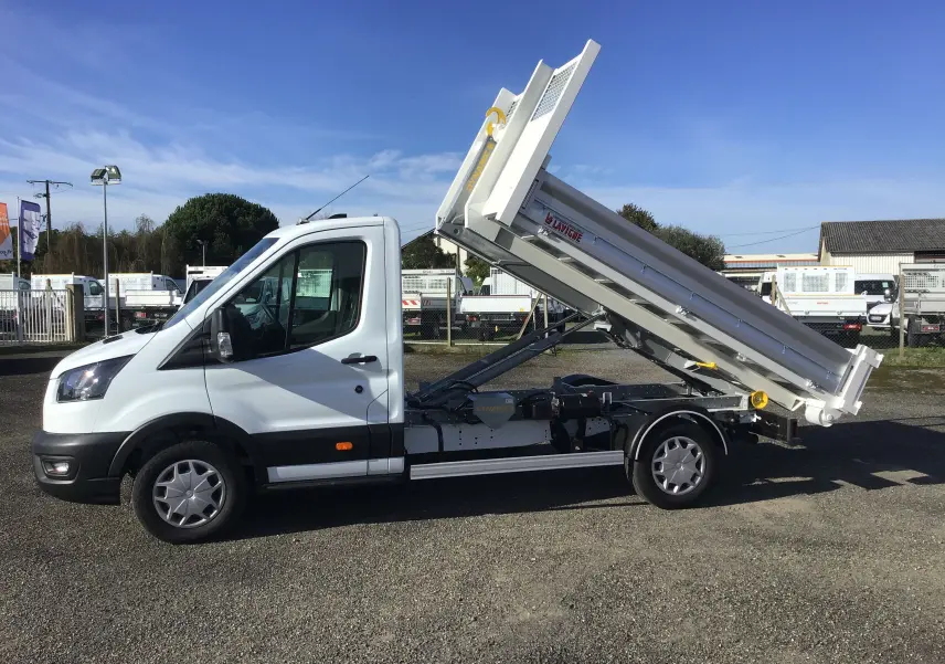 Vue de profil côté gauche d'un Ford Transit blanc avec benne basculante relevée sur un parking extérieur.