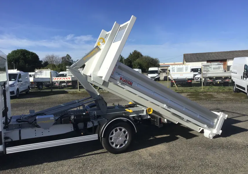 Vue latérale droite d'un Ford Transit blanc avec benne relevée et bras polybenne visible sur un terrain extérieur.
