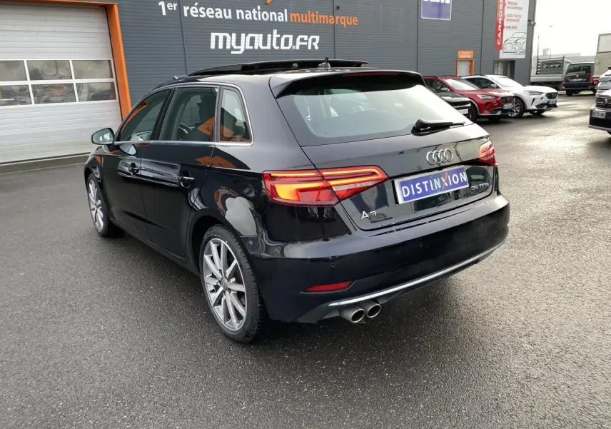 Audi A3 Sportback noire vue 3/4 arrière droit, feux arrière LED allumés et double sortie d’échappement chrome.