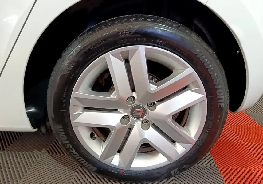 Gros plan sur la roue arrière droite d'une Renault Clio blanche, avec passage de roue visible et sol en damier rouge et gris.