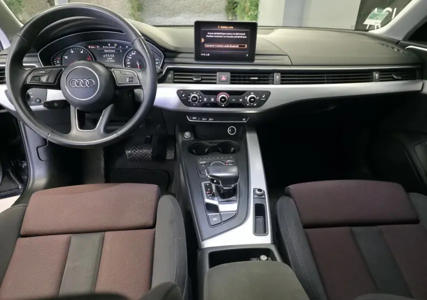 Vue intérieure avant de l'Audi A4 2019, tableau de bord noir avec écran central et console argentée, sièges tissu marron.