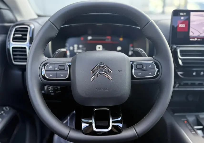 Vue rapprochée du volant noir en cuir du Citroën C5 Aircross 2025 avec commandes intégrées et tableau de bord numérique flou.