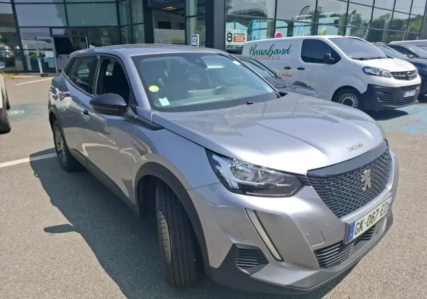 Peugeot 2008 gris Artense en 3/4 avant droit, avec calandre noire et jantes alliage foncé, stationnée sur un sol gravillonné.