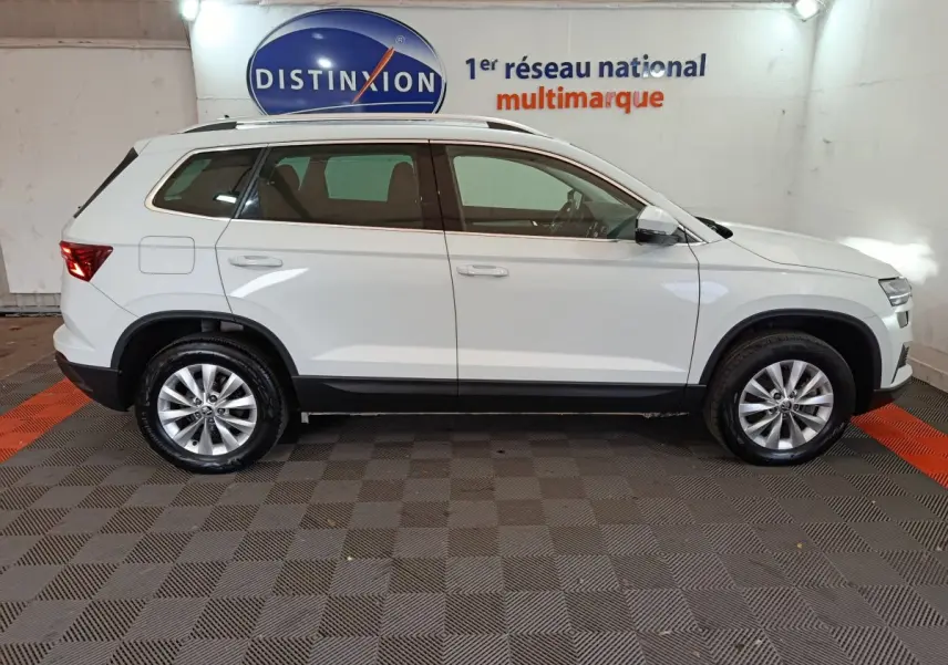 Vue latérale droite d'un SKODA KAROQ blanc 2024 garé en intérieur avec jantes alliage et bas de caisse noir.