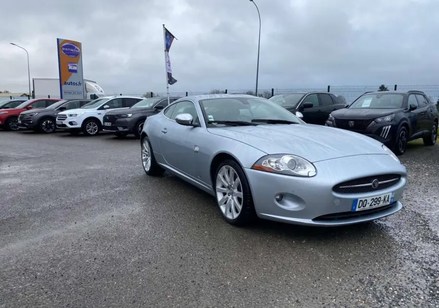 Jaguar XK Coupé gris clair vu en 3/4 avant droit, avec ses jantes alliage et calandre ovale distinctive.