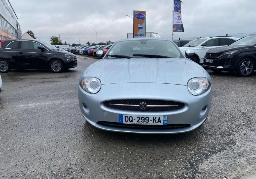 Jaguar XK Coupé gris clair vu de face sur un parking sous un ciel nuageux avec des gouttes de pluie visibles.