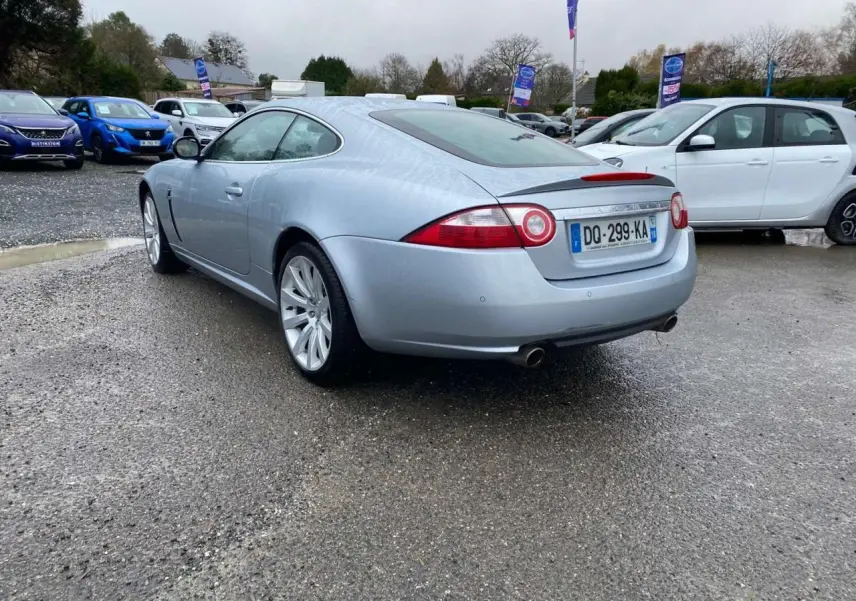 Jaguar XK Coupé gris clair vue 3/4 arrière droit, avec feux ronds caractéristiques et double sortie d'échappement.