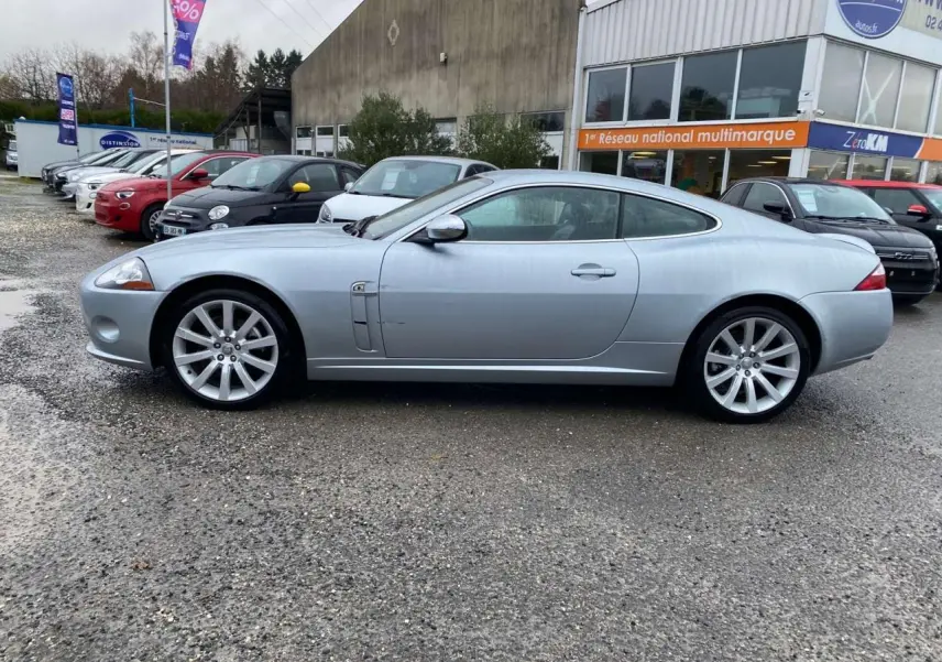 Jaguar XK Coupé gris clair vu de profil côté gauche, stationné devant un bâtiment commercial.