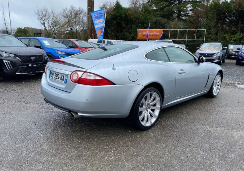 Jaguar XK Coupé gris clair vu 3/4 arrière droit, avec jantes alliage et feux arrière ronds distinctifs.