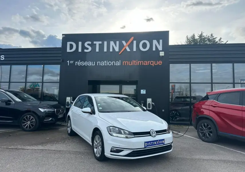 Volkswagen Golf VII blanche vue 3/4 avant droit, stationnée devant un showroom Distinxion avec borne de recharge.