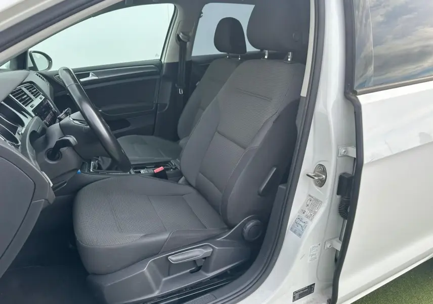 Vue intérieure côté conducteur de la Volkswagen Golf VII blanche, montrant les sièges avant en tissu gris et le volant.