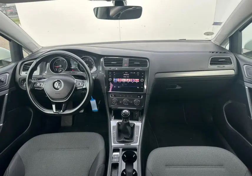 Vue intérieure avant de la Volkswagen Golf VII blanche, tableau de bord noir avec console centrale et volant cuir multifonction.