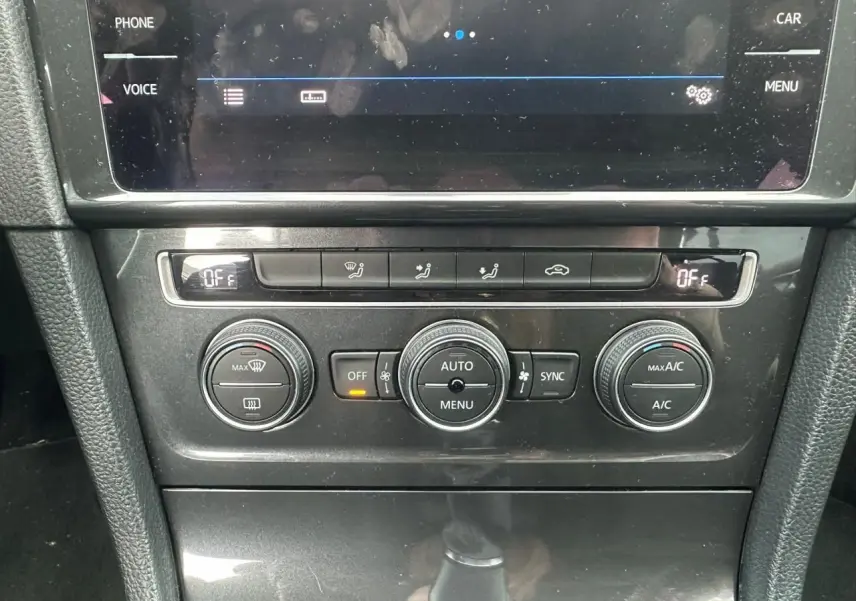 Gros plan sur la console centrale noire de la Volkswagen Golf VII blanche, affichant les commandes de climatisation automatique.