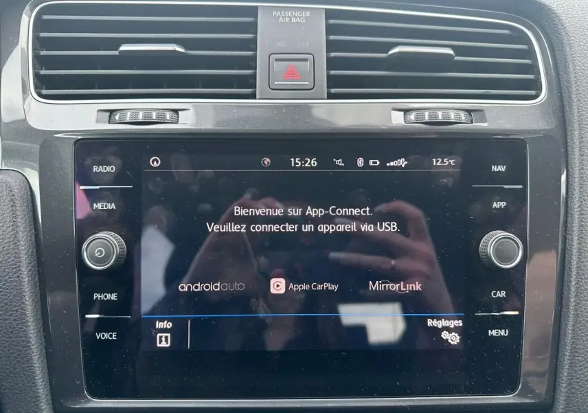 Gros plan sur l'écran tactile central noir de la Volkswagen Golf VII blanc, affichant App-Connect et commandes autour.
