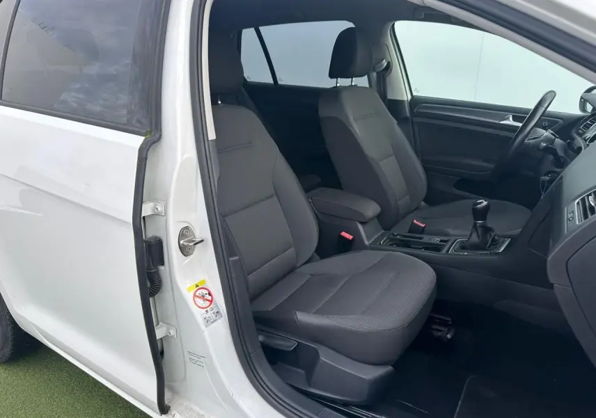 Vue intérieure côté gauche avant d'une Volkswagen Golf blanche avec sièges tissu gris et levier de vitesse manuel.
