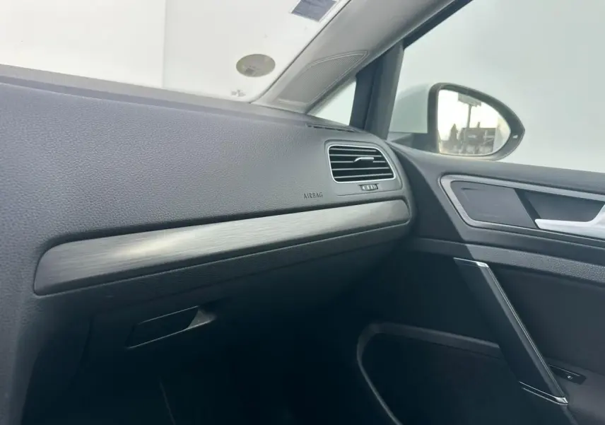 Vue intérieure côté passager avant de la Volkswagen Golf VII blanche, soulignant la planche de bord avec insert décoratif métal brossé.