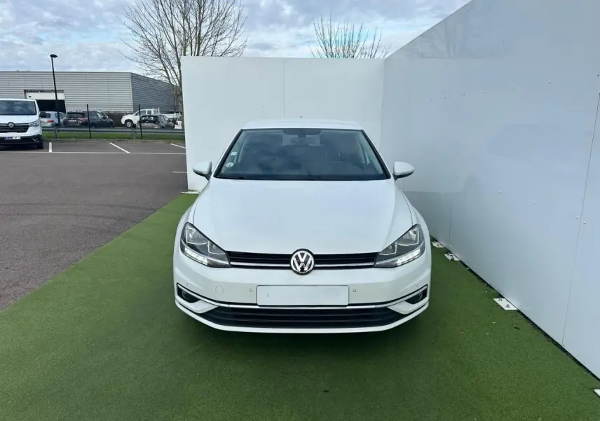 Vue frontale d'une Volkswagen Golf VII blanche garée sur un sol vert, avec calandre noire et logo VW visible.