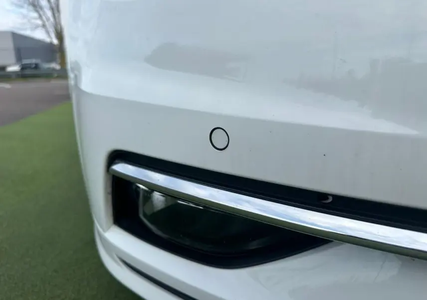 Gros plan sur le pare-chocs avant blanc d'une Volkswagen Golf VII avec capteur de stationnement et insert chromé.