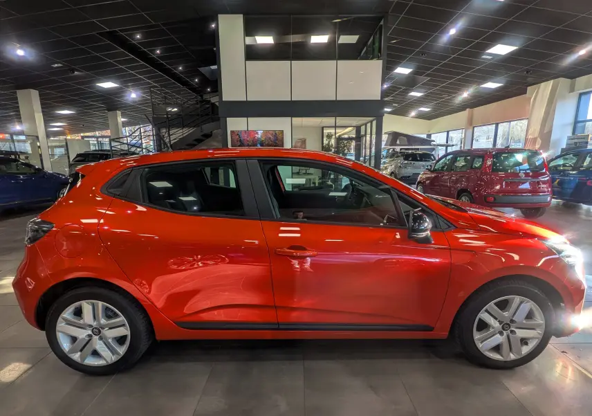 Profil côté gauche d'une Renault Clio V Orange Valencia 2024, compacte et brillante en intérieur de showroom.