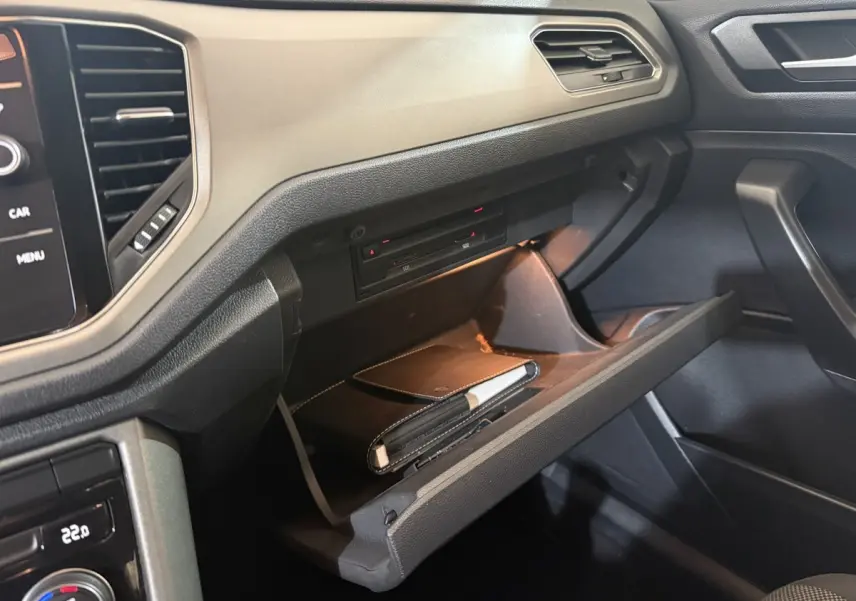 Vue rapprochée de la boîte à gants ouverte du Volkswagen T-Roc noir, mettant en valeur son intérieur et rangement éclairé.